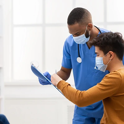 Mann in blauem Kittel und medizinischem Mundschutz zeigt einem weiteren Mann mit Mundschutz Informationen auf einem Klemmbrett